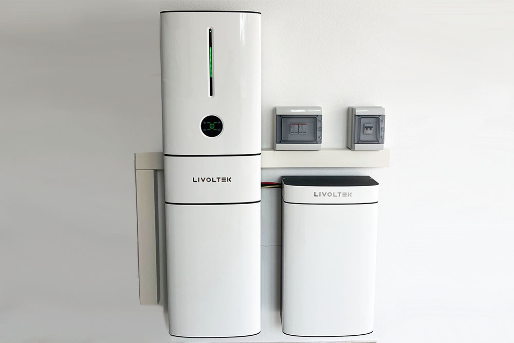 Primo piano di un'installazione domestica a parete di un sistema di accumulo residenziale del marchio Livoltek. Sono visibili l'inverter stack e un pacco batteria aggiuntivo di colore bianco, montati su una parete chiara. Sopra le unità, sono presenti due quadri elettrici grigi di protezione e canaline bianche. Il sistema mostra un indicatore LED verde acceso sull'unità principale. Nell'angolo in alto a destra è presente il logo 'energisolar SARDEGNA'. L'immagine rappresenta una soluzione di accumulo fisico fotovoltaico in Sardegna per massimizzare l'autoconsumo dopo la fine dello Scambio sul Posto.