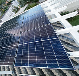 Vista dall'alto di moduli fotovoltaici installati in serie sulle travi di una pergola in legno bianco, con un pavimento a piastrelle esagonali visibile in basso.